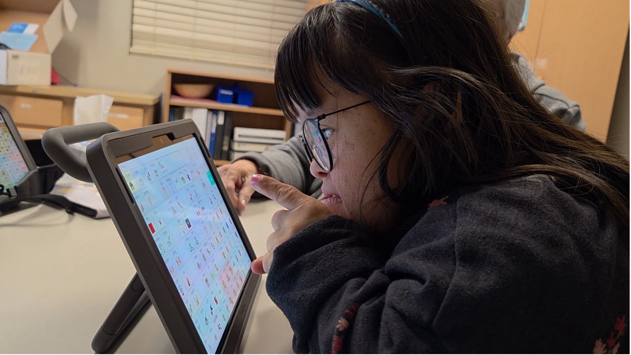 A woman uses her finger to select an icon on her AAC communication device, a tablet displaying a grid of colorful symbol icons