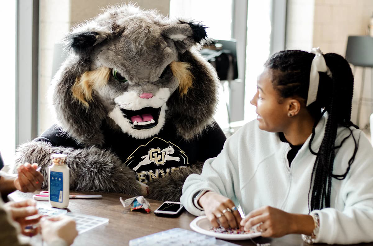 Milo the Lynx and a student seated at a table making friendship bracelets.
