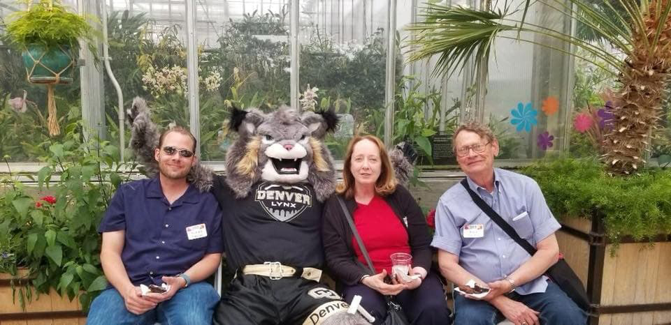 Three people seated with Milo the Lynx mascot surrounded by green foliage.