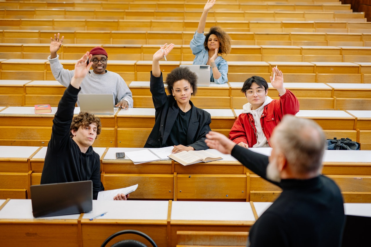 Five students sit in a lecture hall while all of them raise their hands in front of a professor who points his arm toward one of them.