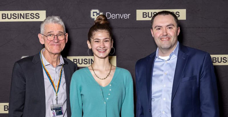 Three people stand together in business professional clothing, posing and smiling against a CU Denver Business School backdrop.