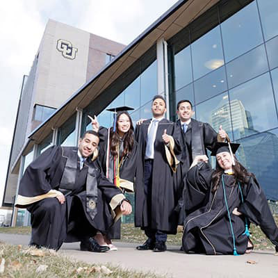 Group of CU Denver students in commencement regalia outside a CU building celebrating together.