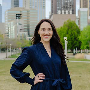 Katie Imhoff stands outside in a long navy dress and long hair, smiling with her teeth. The Denver city scape is in the background.