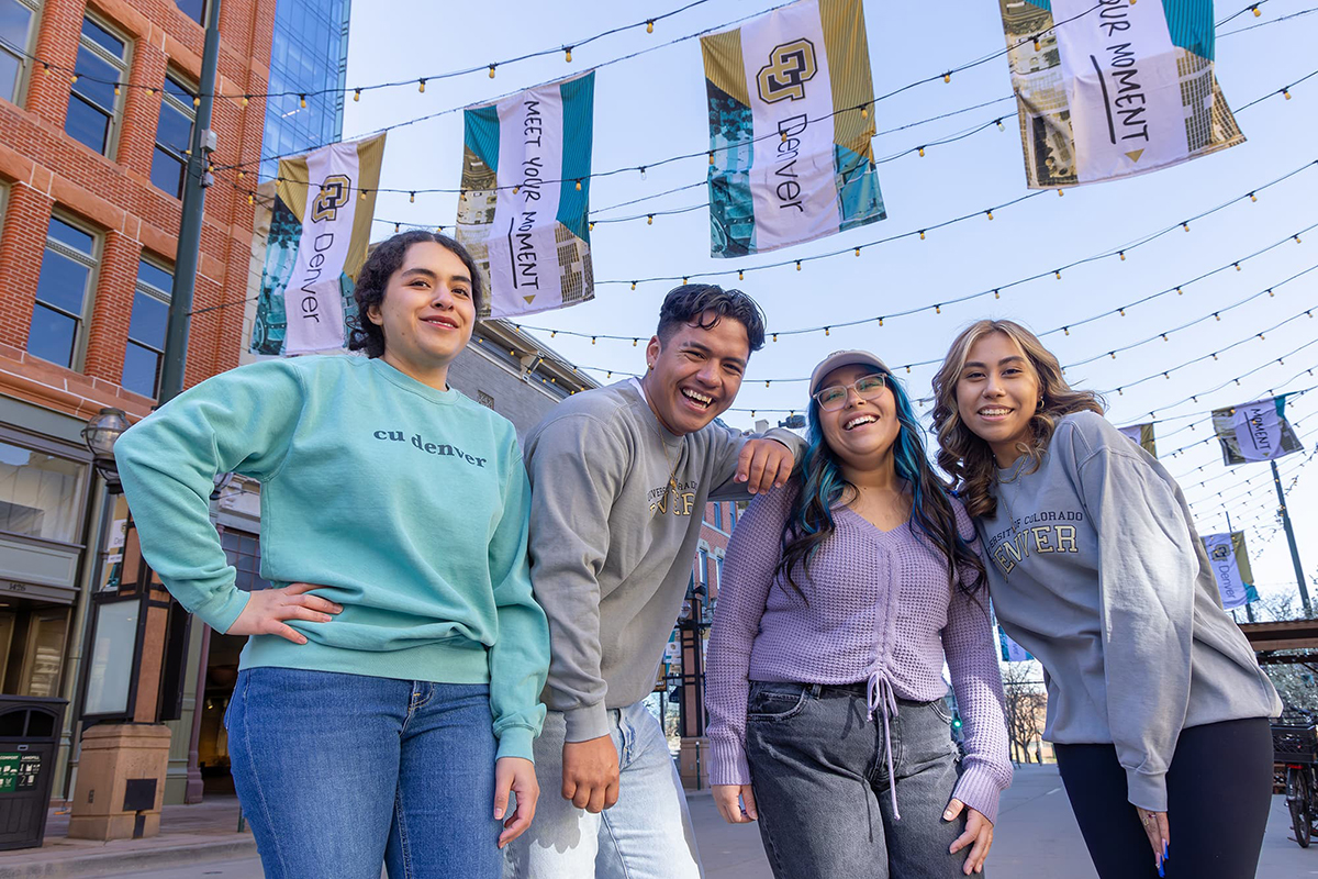 Four people in CU Denver branded gear smiling in a street with CU Denver flags and bistro lights hanging above them.