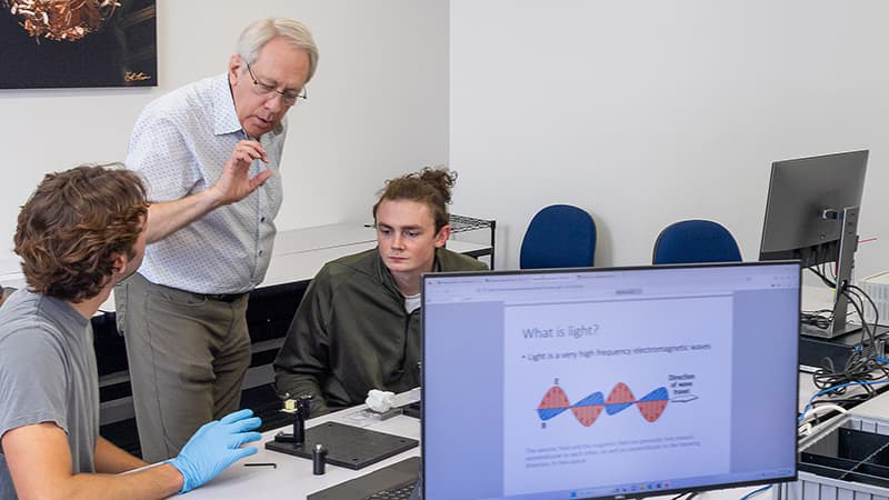 Man in button up shirt, glasses, and short white hair speaks with his hand while two students listen.