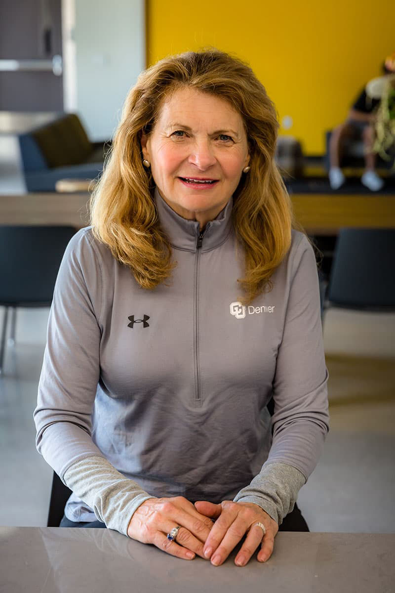 Joanne Posner-Mayer wears a gray CU Denver-branded jacket, long blonde hair, and smiling in her family-named kitchen.