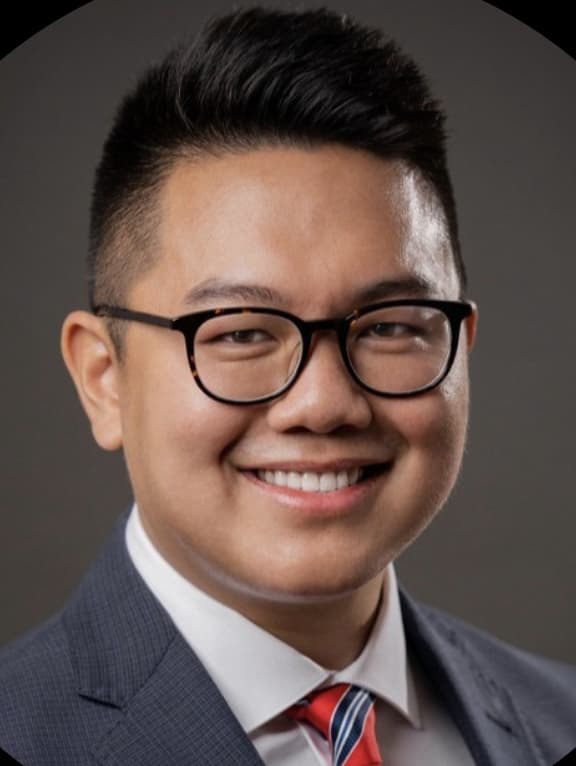 Scott Cao wearing a suit and tie with dark-rimmed glasses, smiling with his teeth.