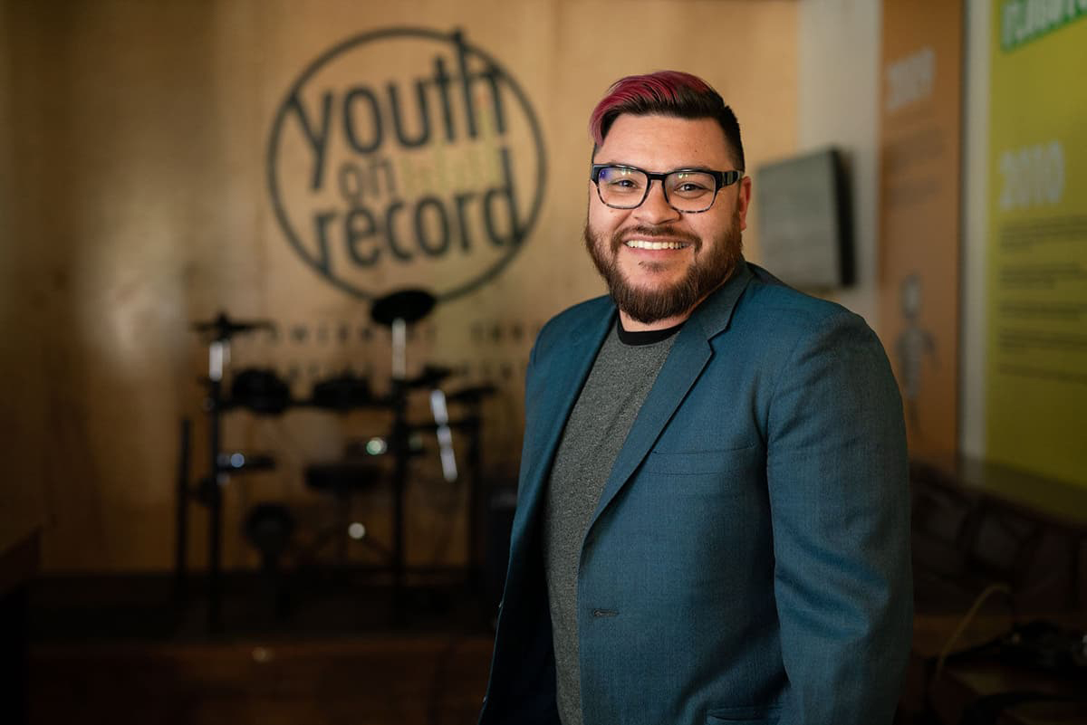 Andy Guerrero in a dark blue blazer, dark, squared-rimmed glasses, and red hair smiles with his teeth.