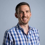 Rob Carpenter in a blue checkered shirt and short brown hair. He has some short, brown facial hair and smiles warmly showing his teeth.