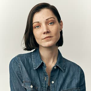 A woman with short, dark hair and wearing a jean button-up blouse looks into the camera with a straight face.