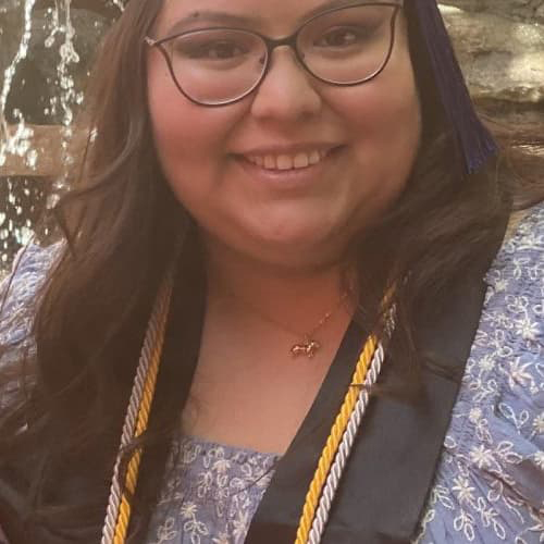 Woman with long hair and dark framed glasses, smiling with her teeth wearing a commencement stole and cords.