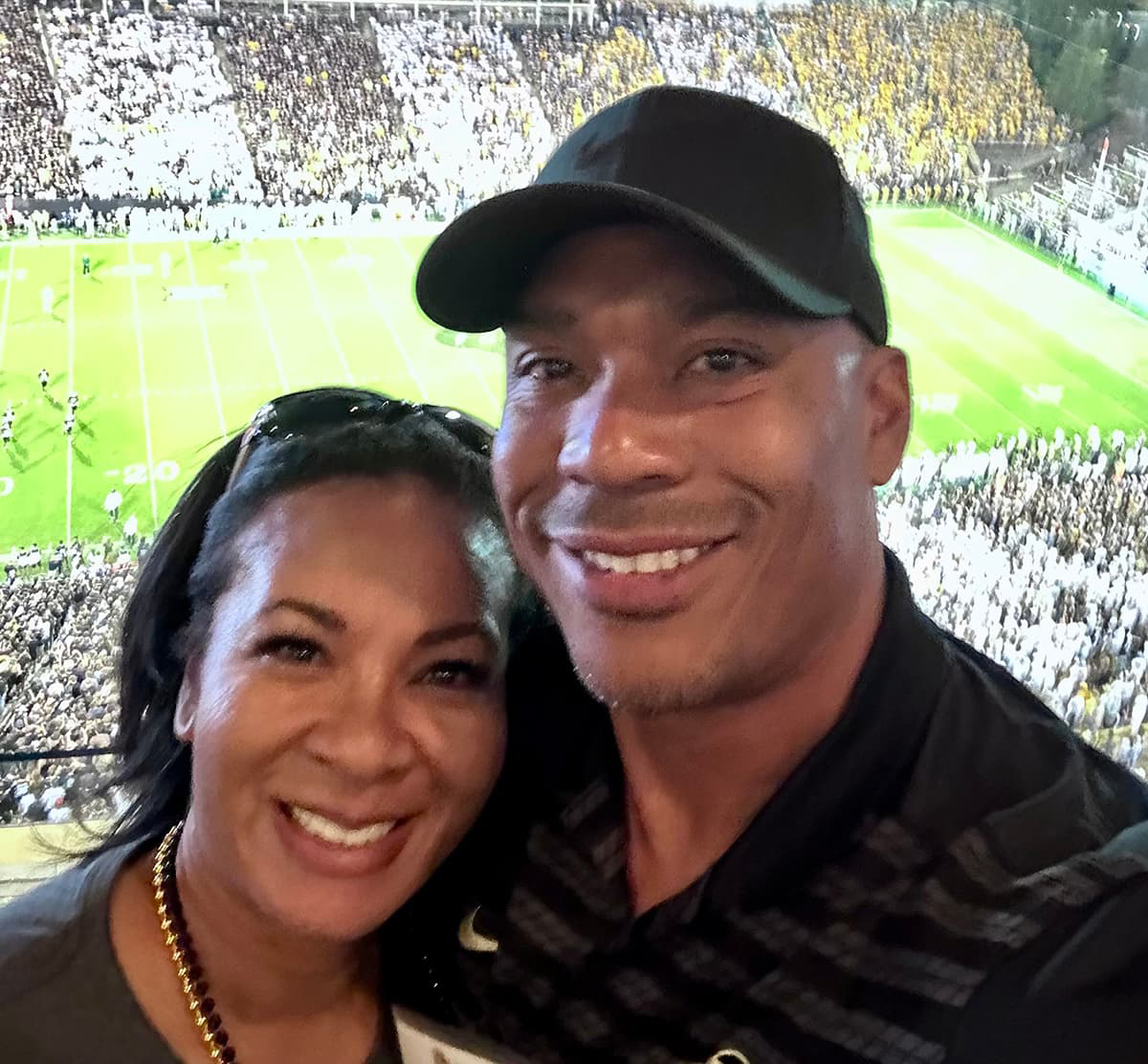 A male and female couple dressed in black taking a selfie with a football game happening behind them.