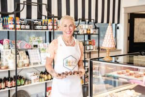 Owner Gretchen Harrison inside Alice   Kate holding a tray of truffles wearing a white apron with short blond hair, smiling with her teeth.