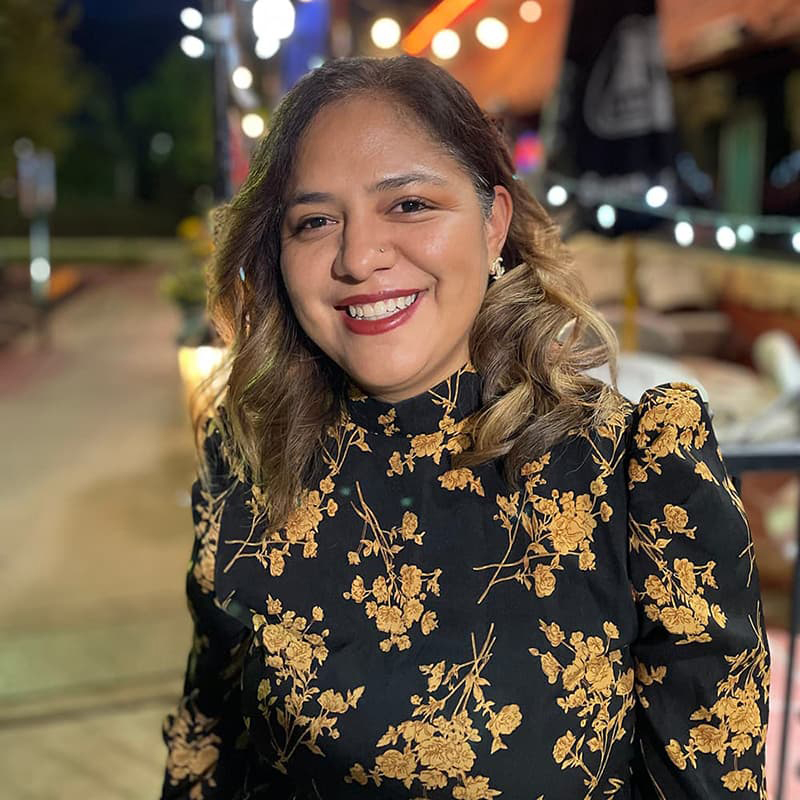 Vanessa Medina Pasillas dressed in a black blouse with gold flowers, mid-length brown-blonde hair and smiling with her teeth.