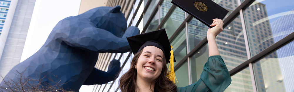 CU Denver graduate holding diploma sleeve up standing in front of Colorado Convention Center